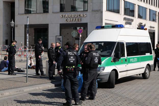 Eine Gruppe von Polizeibeamten steht vor einem Starbucks-Café, rechts davon ein Van und links ein paar Menschen, im Hintergrund sind ein Gebäude, ein Schild, ein Laternenpfahl und ein Zaun zu sehen.