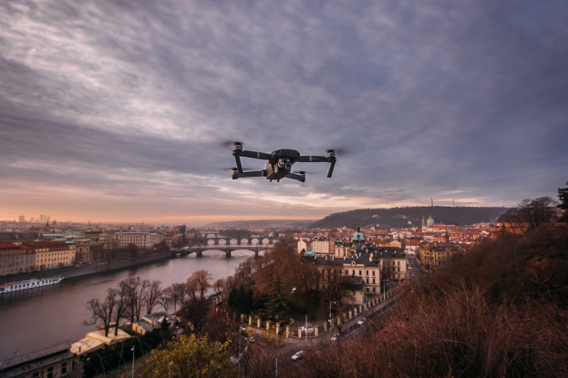 Eine Drohne fliegt über eine Stadt mit Gebäuden, Bäumen und Fahrzeugen, wobei ein Fluss und eine Brücke im Hintergrund zu sehen sind.