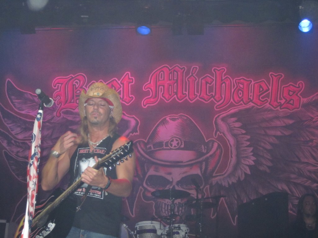 Eine Musik-Konzertszene mit einem Gitarristen links neben einem Mikrofon, einer Person in der unteren rechten Ecke, einem Banner im Hintergrund und Drogen in der Mitte unten, alles unter Bühnenbeleuchtung von oben.