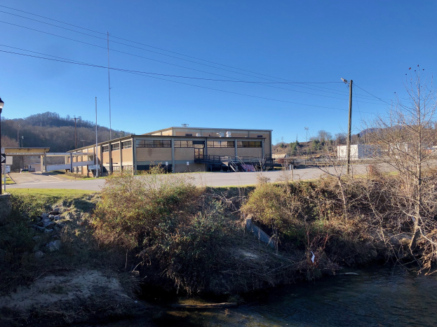 Ein Fluss fließt durch ein grünes Feld neben einem Gebäude neben Bäumen, Pflanzen, Gras, Steinen, Straßenlaternen, Strommasten, Stromkabeln, Hügeln und einem klaren blauen Himmel, wobei das Gebäude für eine Wasseraufbereitungsanlage geplant ist.