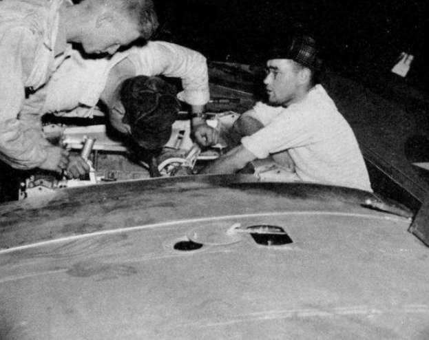 Drei Männer mit Mützen reparieren ein schwarz-weißes Auto in einer Garage, umgeben von Werkzeugen und Materialien.