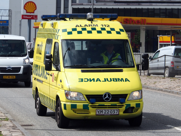 Ein Rettungswagen mit Personen darin fährt auf einer Straße neben einer Tankstelle, mit einem Gehweg auf der linken Seite und Gebäuden, Polen, Schildern, Bäumen und einem klaren blauen Himmel im Hintergrund.