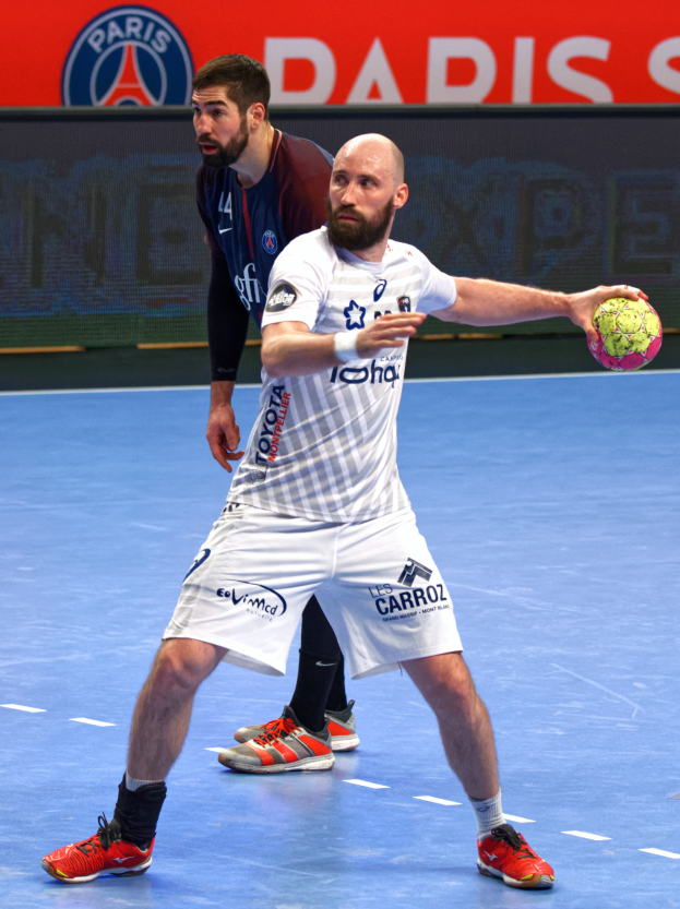 Zwei Männer beim Handballspielen auf einem Platz, einer hält den Ball, im Hintergrund eine Tafel mit 'Paris Saint-Germain vs Paris Saint Germain'.