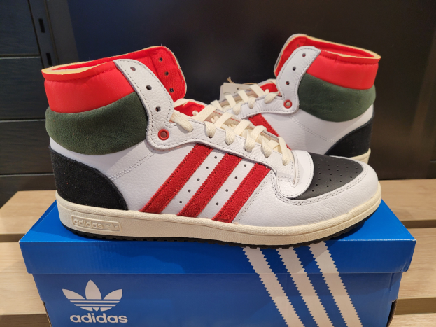 Ein Paar weiße, rote, grüne und schwarze Adidas Originals Top Ten Hi-Schuhe auf einer blauen und braunen Kartonschachtel, die auf einem cremefarbenen Tisch mit einer schwarzen Wand im Hintergrund steht.