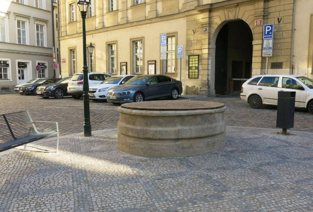 Ein Stadtplatz mit einer Bank, einem Brunnen, Gebäuden, Laternen und Schildern.