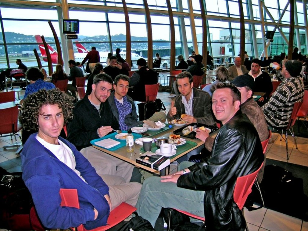 Eine Gruppe von Menschen sitzt an einem Tisch mit roten Stühlen in einem Flughafen, isst und unterhält sich, mit einer Glaswand, einem Hügel, einem Flugzeug und einem Fernseher im Hintergrund.