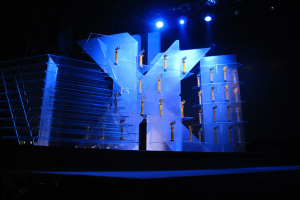 Eine Bühne, die für eine Preisverleihung aufgebaut ist, mit einem dunklen Hintergrund, der von ein paar Lichtern beleuchtet wird, mit Gegenständen, die wahrscheinlich mit dem Ereignis zu tun haben.