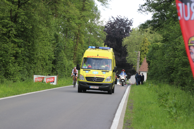 Ein Rettungswagen fährt auf einer Straße mit Radfahrern daneben, umgeben von Gras, Bäumen, Häusern, Pfählen und einem klaren blauen Himmel.