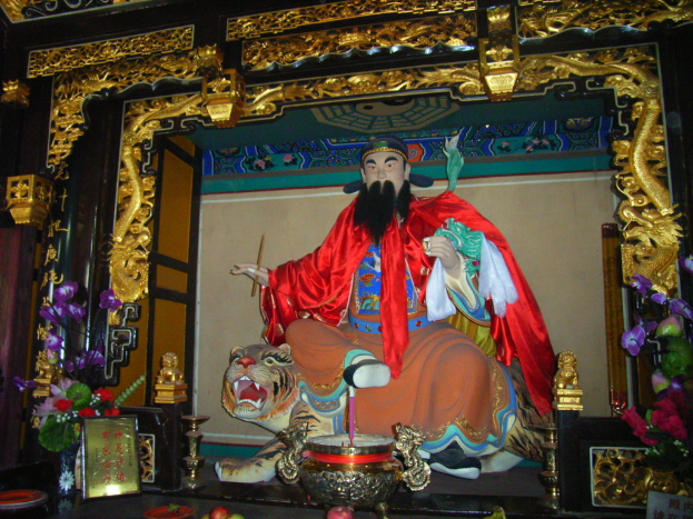 Eine Statue eines chinesischen Mönchs, der mit buntem Stoff geschmückt ist, sitzt auf einer Tigerstatue innerhalb eines Bogens mit goldenen Verzierungen, vor ihm steht ein Blumenkübel und in der Mitte eine Schüssel.