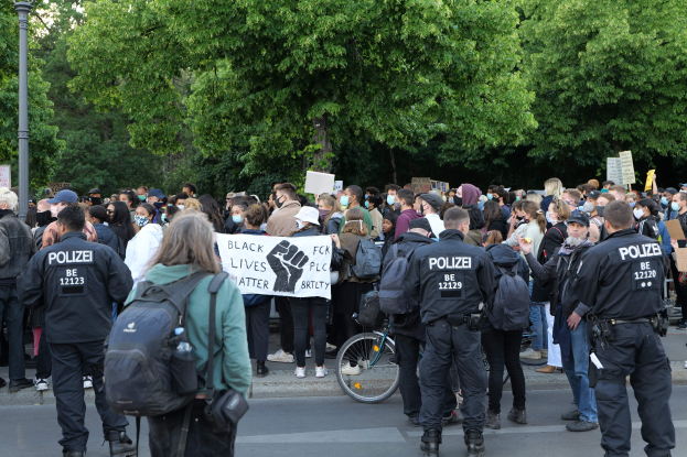 Eine große Gruppe von Menschen bei einer Black Lives Matter Demonstration in Berlin, einige halten Schilder und tragen Mützen und Taschen, mit einem Fahrrad im Vordergrund und Bäumen und einem Mast im Hintergrund.