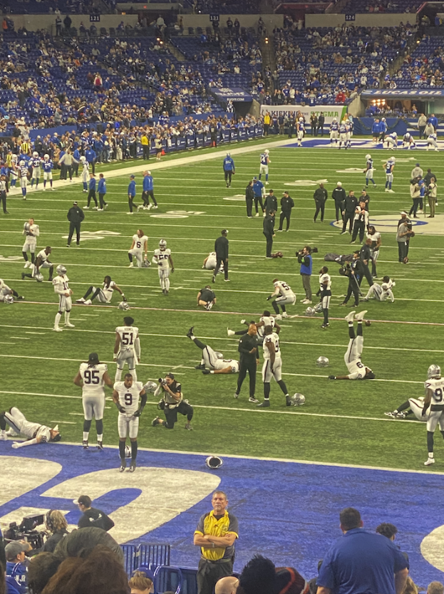 Ein Fußballspiel wird in einem Stadion mit Zuschauern und einigen Sitzenden sowie einer Person mit einer Kamera gespielt.
