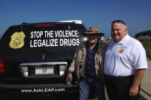 Zwei Männer stehen neben einem schwarzen Van mit "Stop the Violence Legalize Drugs" darauf, einer hält eine Flasche, mit Menschen, Bäumen und einem klaren blauen Himmel im Hintergrund.