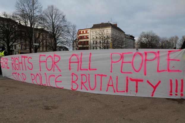 Eine Gruppe von Menschen, die auf dem Boden stehen und ein Banner halten, auf dem "Rechte für alle Menschen - Stoppt Polizeigewalt" steht, mit einer Straßenlaterne, einem Schild, Bäumen, Gebäuden mit Fenstern und einem bewölkten Himmel im Hintergrund.