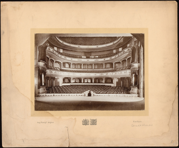 Altes Schwarz-Weiß-Foto des Auditoriums des Royal Albert Hall, das die Bühne, Reihen von Stühlen, Säulen, Geländer und Text mit einem Logo unten zeigt.