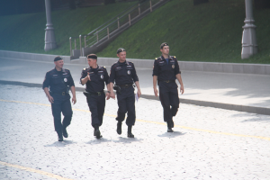 Drei Polizisten in Uniform und mit Mützen, die eine Straße mit Pfählen, Geländern, Treppen und Gras im Hintergrund entlanggehen.