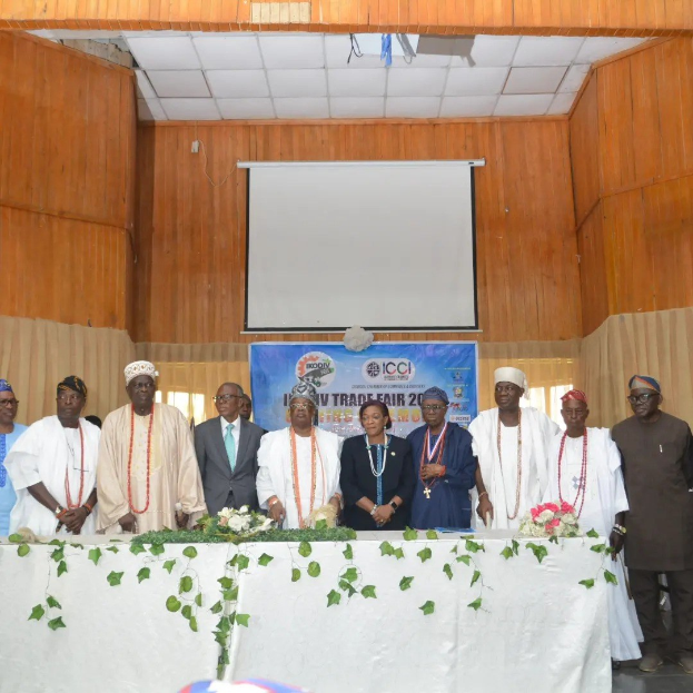 Gruppe von Menschen um einen weißgedeckten Tisch versammelt, der mit Blumen und Blättern geschmückt ist, mit einem "ICCI International Trade Fair 2019"-Banner, Projektionsbildschirm und Deckenbeleuchtung im Hintergrund.