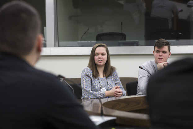 Eine Gruppe von Menschen sitzt um einen Tisch mit Papieren und einem Mikrofon vor einem Glasfenster und ist in eine Diskussion vertieft.