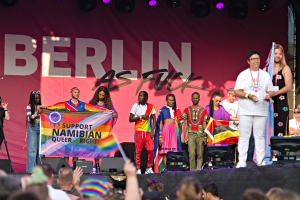 Gruppe von Menschen auf einer Bühne mit einem Banner, auf dem "Berlin as Fuck" steht, mit Lautsprechern, Mikrofonen und einer feiernden Menge unten, die Hände hebt.