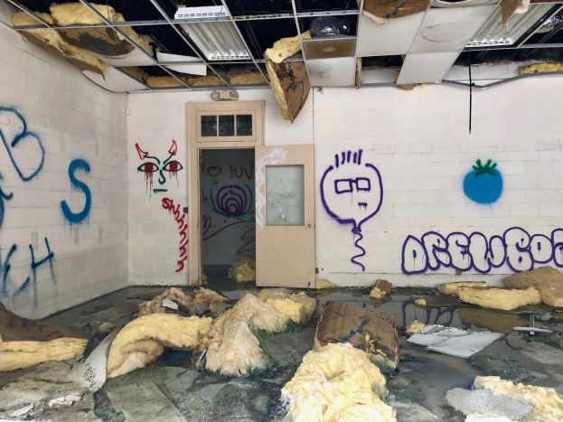 Ein Raum mit Graffiti an den Wänden, eine Tür im Hintergrund, Deckenstangen und Lampen sowie verschiedene Gegenstände auf dem Boden.