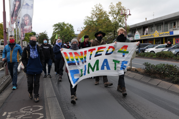 Eine Gruppe von Menschen marschiert mit einem "United 4 Climate"-Schild die Straße entlang, trägt Masken und trägt Taschen, mit einem Gehweg, Geländer und Schildern auf der linken Seite, Bäumen, Laternenmasten, Fahrzeugen, Gebäuden und einem klaren blauen Himmel im Hintergrund.