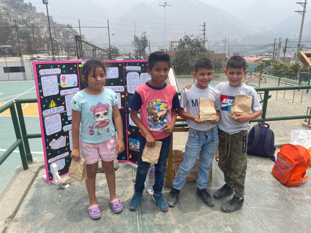 Eine Gruppe von Kindern steht auf einem Basketballplatz, jedes hält einen Papiertüte, mit Tüten und Gegenständen rechts und einer Tafel mit Text links, mit Zaun, Pfosten, Straßenlaternen, Strommasten mit Drähten, Bäumen, Gebäuden, Hügeln und dem Himmel im Hintergrund.