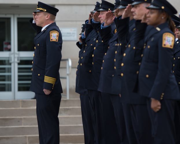 Eine Gruppe von Polizisten in Uniform steht in Formation, salutiert vor einem Gebäude mit Glastüren und Treppen.
