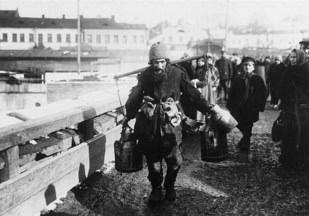 Eine Gruppe von Menschen, die wie Flüchtlinge aussehen, geht eine Straße entlang neben einem Gewässer, mit einem hölzernen Zaun links und Gebäuden und Masten im Hintergrund, dargestellt in Schwarz-Weiß.