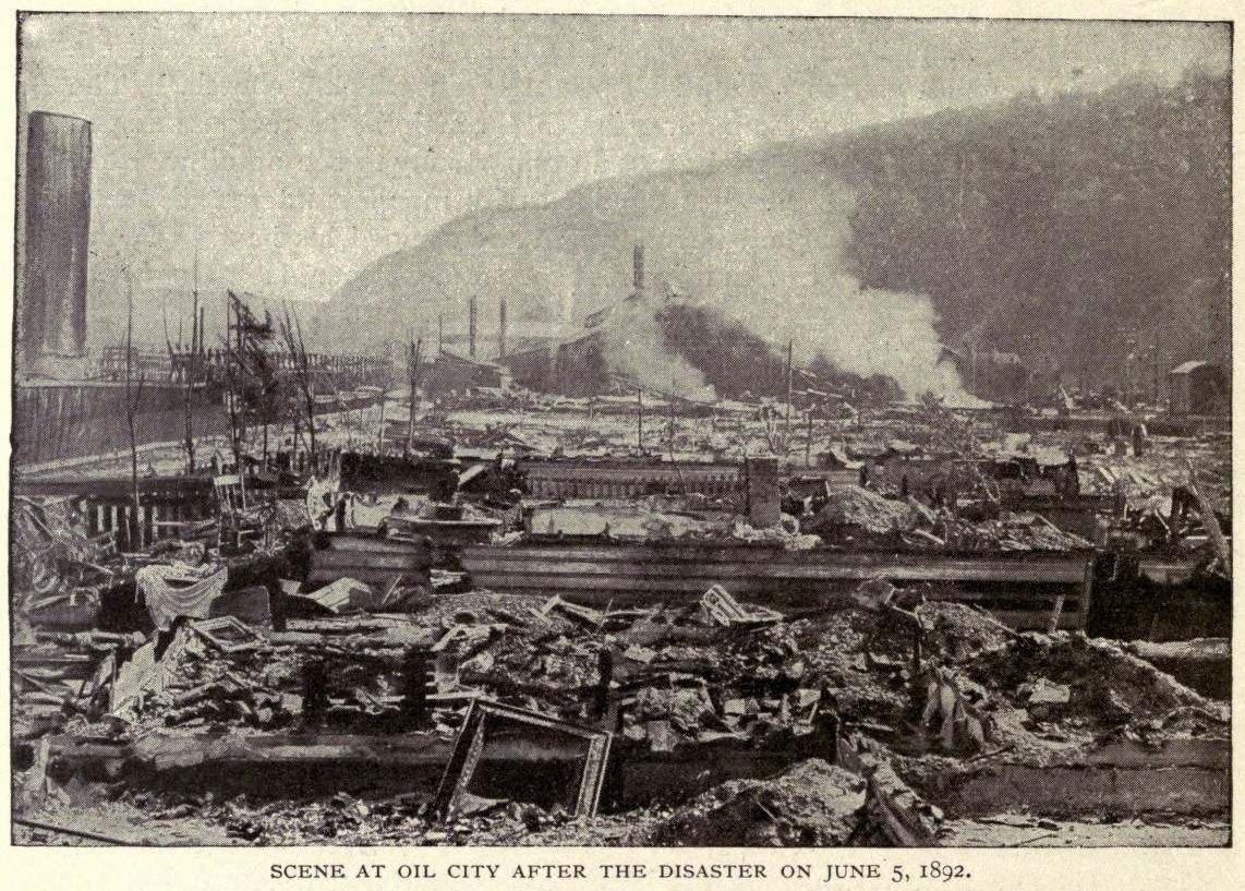 Schwarzes und weißes Foto einer Stadt nach einer Katastrophe am 5. Juni 1892, das beschädigte Gebäude, Strommasten, Rauch und einen Hügel im Hintergrund zeigt, mit Text unten.