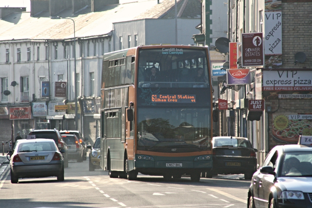 Eine Straße mit Autos und einem Bus vor Gebäuden mit Wänden, Fenstern, Tellern und Dächern, mit Plakaten und Bannern an den Wänden und einem Mast mit einer Straßenlaterne.
