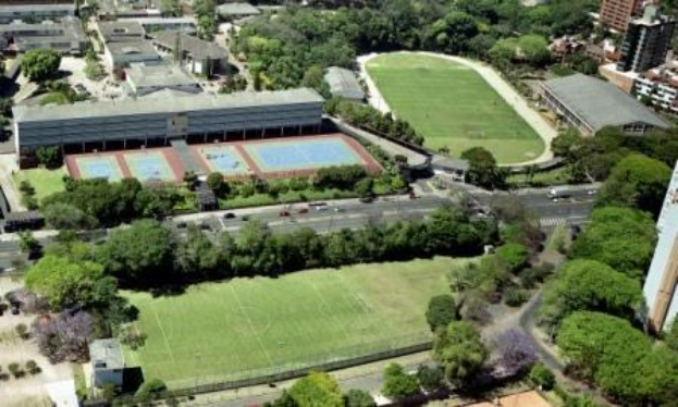 Luftaufnahme eines Fußballfeldes auf dem Campus der Universität Sydney, umgeben von Bäumen, Gebäuden und Fahrzeugen, mit einem neuen Sportkomplex in Entwicklung.
