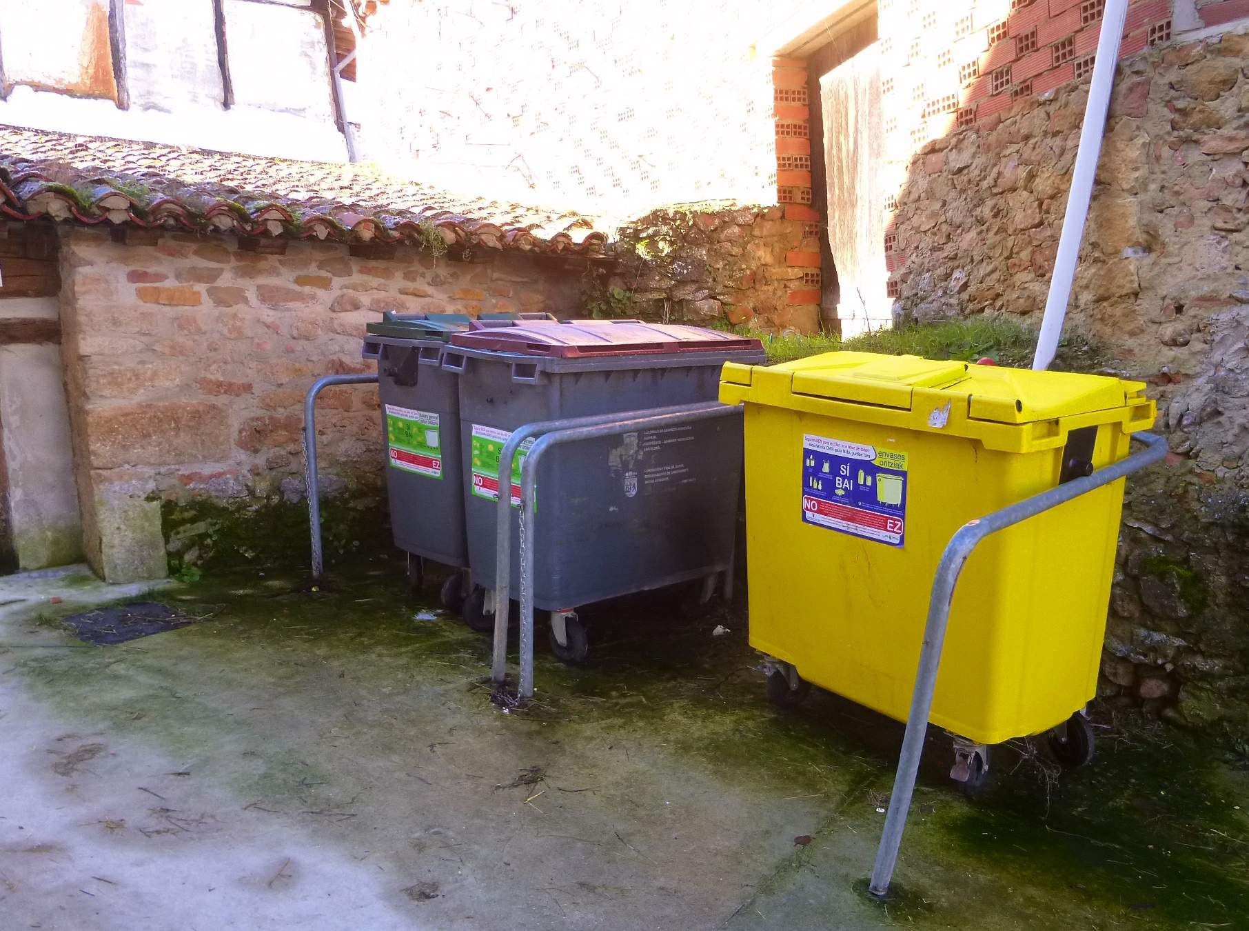 Zwei gelbe und graue Recyclingcontainer mit Rädern nebeneinander vor einem Gebäude mit einer Wand und einem Fenster positioniert.