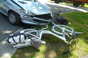 Ein verunglücktes Auto am Straßenrand mit einem Fahrrad auf dem Gras daneben, Bäume im Hintergrund unter einem klaren blauen Himmel.