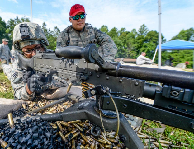 Eine Person auf einem Armee-Trainingsgelände feuert eine Waffe ab, neben ihr steht eine Person mit roter Mütze, während im Hintergrund Bäume, Zelte und Menschen unter einem klaren, sonnigen Himmel zu sehen sind, und im Vordergrund Patronenhülsen und Gras liegen.