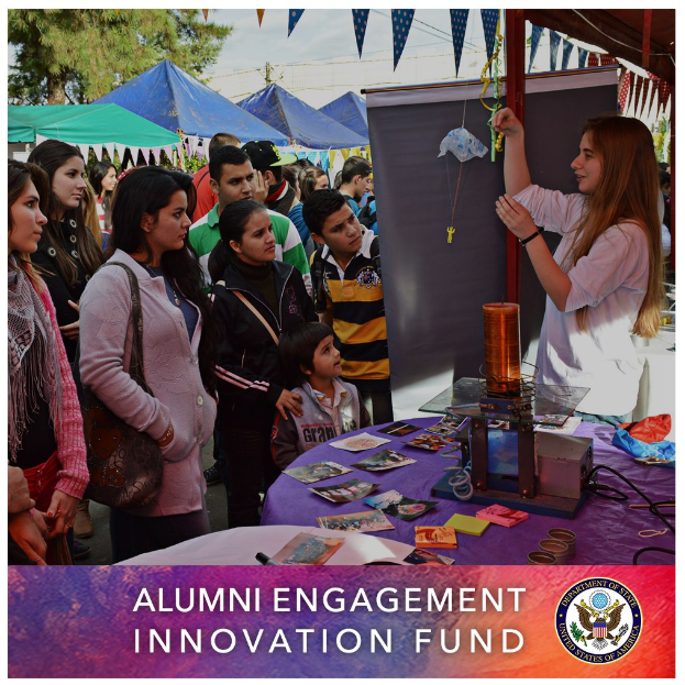 Eine Gruppe von Menschen um einen Tisch herum mit einer 'Alumni Engagement Innovation Fund'-Tafel, mit Gegenständen auf dem Tisch und Zelten, Bäumen und einem klaren blauen Himmel im Hintergrund.