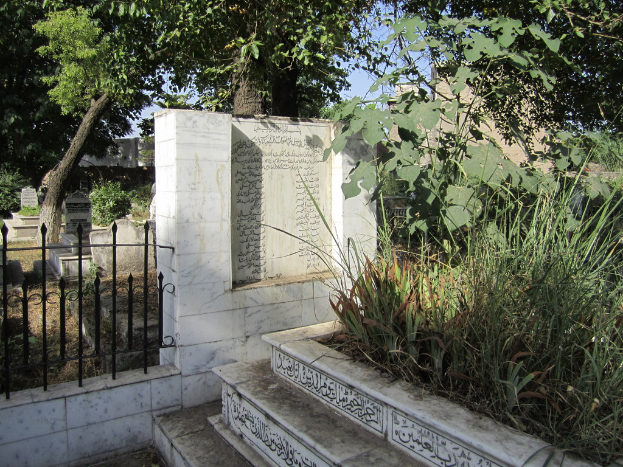 Eine Gedenktafel neben einem Geländer in einem Friedhof, mit Treppen unten, Gras und Bäumen rechts, Gräbern hinter dem Geländer und Gebäuden und Bäumen im Hintergrund.