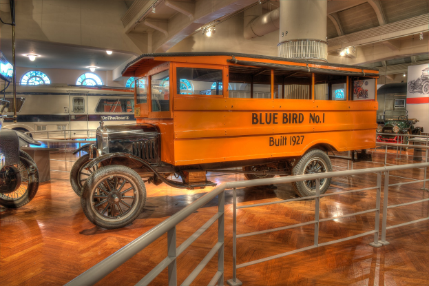 Ein orangefarbener Bus in einem Museum ausgestellt, umgeben von anderen Fahrzeugen, mit einem Geländer davor und einem Rad im Vordergrund.