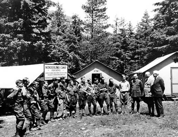 Eine Gruppe von Männern in einem Schwarz-Weiß-Foto, die vor einem Zelt mit Bäumen und einem Schild im Hintergrund stehen und als Mitglieder des Wonderland Camp für Jungs identifiziert werden.