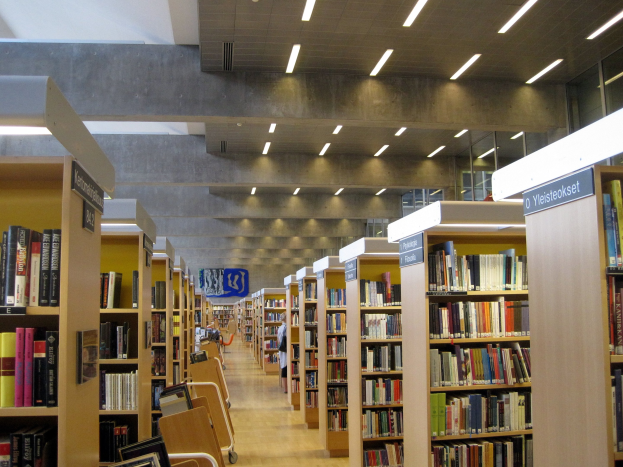 Eine geräumige Bibliothek mit hohen Bücherregalen, Stühlen, Informationsplakaten, Deckenleuchten und Glasfenstern.