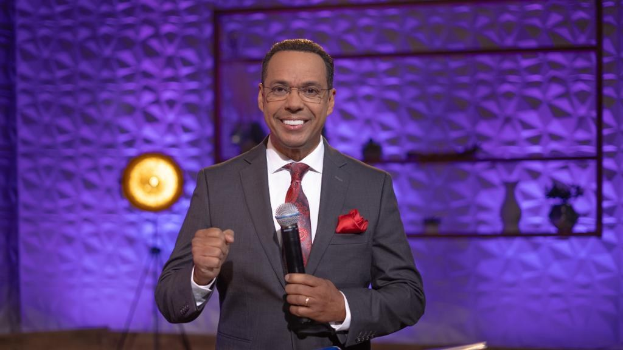 Stephen Colbert, Moderator von "The Daily Show with Trevor Noah", steht auf einer Buhne und halt einen Mikrofon in der Hand, tragt einen Anzug, Krawatte und Brille, mit Licht- und anderen Geraten im Hintergrund.