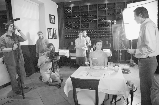 Ein Filmset mit einem Mann auf der rechten Seite, einer Frau an einem Tisch mit Tellern, Gabeln und Gläsern in der Mitte und zwei Personen, die eine Kamera bedienen, auf der linken Seite, mit Gestellen, Rahmen und einem Bildschirm im Hintergrund.