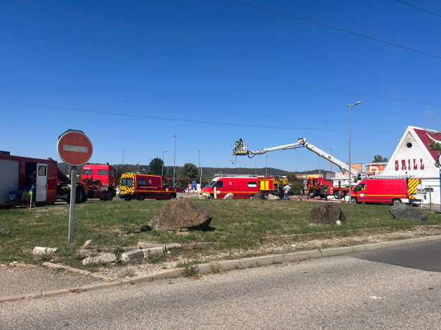 Eine Gruppe von Feuerwehrautos steht auf der Seite einer Straße, umgeben von Gras, Steinen, einem Schild, einem Haus, Masten, einem Kran, Drähten, Bäumen und einem bewölkten Himmel, wahrscheinlich nach einem Feuer an einer Tankstelle.