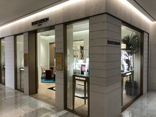 Der Eingang zu einem Boutique in einem Einkaufszentrum, mit Glaswänden und -türen, Text an den Wänden, Deckenleuchten, Stühlen, Tischen, einer Pflanze und anderen Gegenständen, mit dem Boden unten.