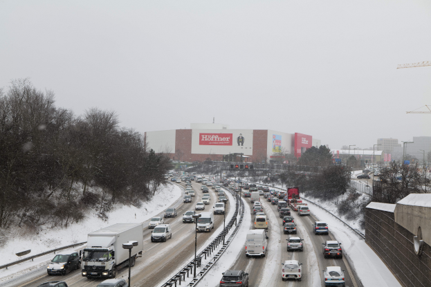 Eine schneebedeckte, stark befahrene Autobahn mit zahlreichen Fahrzeugen, umgeben von Bäumen, Mästen, Lichtern, Schildern, Gebäuden und Kränen, unter einem sichtbaren Himmel.