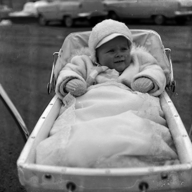 Ein Schwarz-Weiß-Foto eines löchelnden Babys in einem Kinderwagen, das eine weiße Kleidung und eine Kappe trägt, mit Fahrzeugen im Hintergrund.