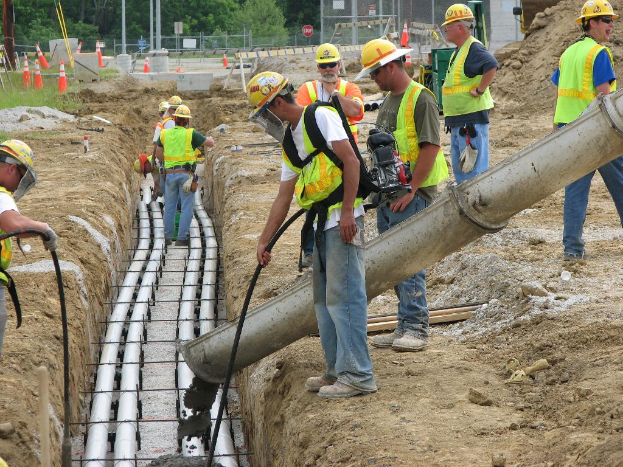 Eine Baustelle mit Arbeitern in Jacken und Helmen, Absperrbaken, Zaun, Bäume, Pfosten, Erde und Rohren im Hintergrund.