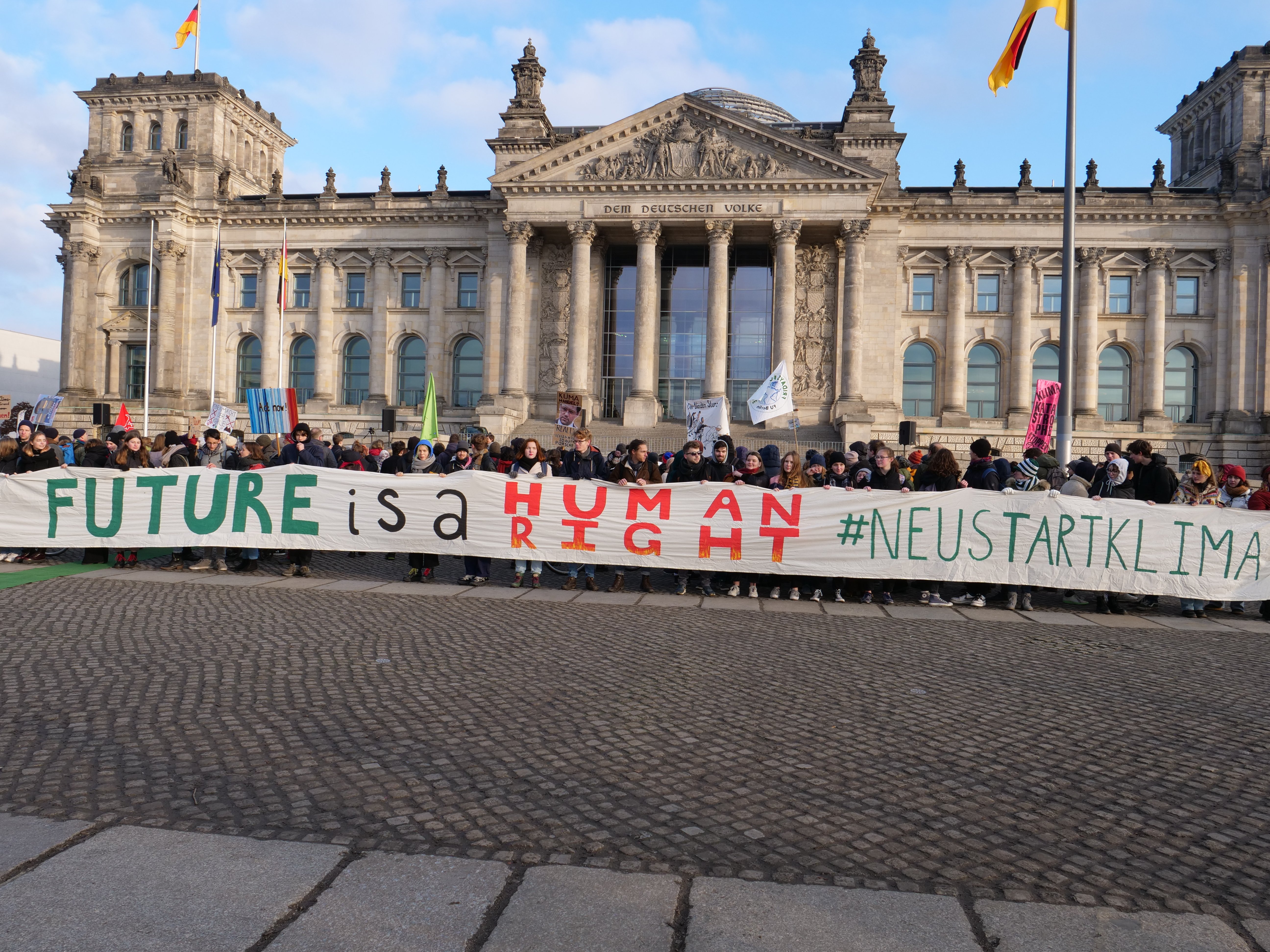 Eine Gruppe von Menschen hält ein Transparent mit der Aufschrift "Zukunft ist ein Mensch" vor dem Reichstaggebäude in Berlin, Deutschland, das mit Fenstern, Säulen, Bögen und Statuen verziert ist und von Fahnenmasten umgeben ist.
