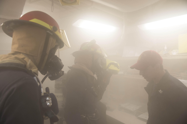 Feuerwehrleute in Helmen und Ausrüstung atmen Rauch während einer Übungseinheit, einer hält eine Kamera, mit Hintergrundbeleuchtung und Equipment.