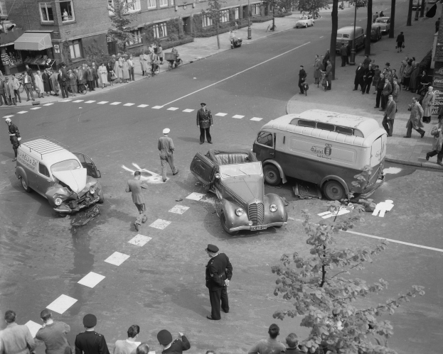 Schwarz-weißes Bild eines Autounfalls an einer Straßenkreuzung mit mehreren Fahrzeugen, darunter ein Van, und einer Gruppe von Menschen in der Nähe, mit Gebäuden, Bäumen, Pfählen und Texttafeln im Hintergrund.