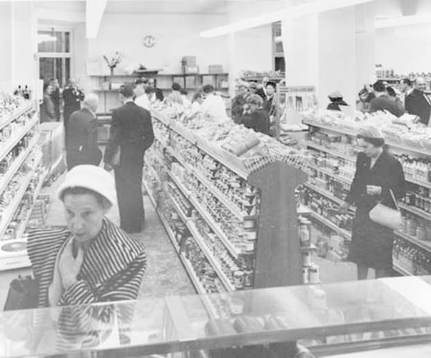Schwarzes und weißes Foto von Menschen, die in einem Supermarkt einkaufen, mit Regalen voller Lebensmittel, einige halten Taschen, eine Uhr an der Wand und Deckenbeleuchtung.