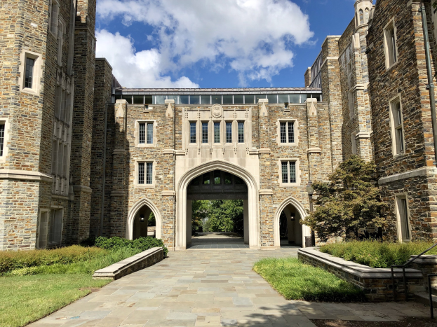 Ein großes steinernes Universitätsgebäude mit einem Weg, umgeben von Grün, mit Fenstern, einem Bogen und einem bewölkten Himmel.